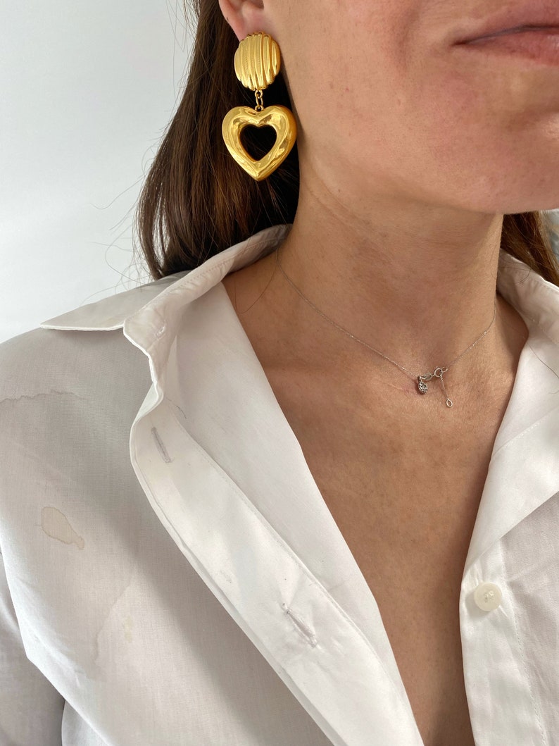 May include: Gold heart-shaped earrings with a textured design. The earrings are dangling and have a smaller, round, textured piece above the heart.