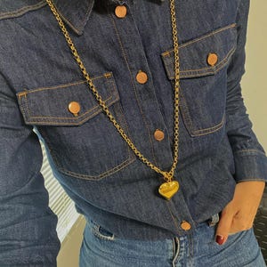 May include: A gold chain necklace with a large heart pendant. The necklace is worn over a denim shirt and jeans.