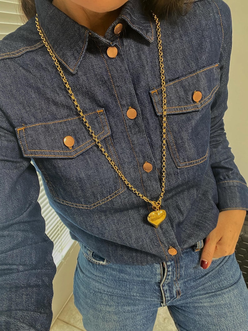 May include: A gold chain necklace with a large heart pendant. The necklace is worn over a denim shirt and jeans.