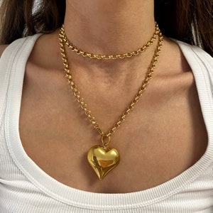 May include: A gold chain necklace with a large, heart-shaped pendant. The chain is layered with a shorter, thinner chain.