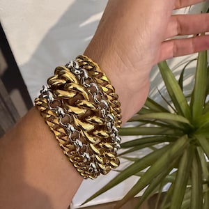 Könnte beinhalten: Ein breites, mehrreihiges Armband mit abwechselnd dicken, goldfarbenen Panzerketten und dünneren, silberfarbenen Gliederketten. Das Armband besteht aus mehreren ineinandergreifenden Metallgliedern, die eine geschichtete, strukturierte Optik erzeugen.