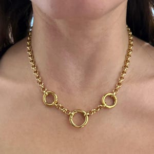 May include: A gold-colored necklace with a chain of small, round links. The necklace features three larger, circular pendants evenly spaced along the chain. The necklace is worn around the neck.