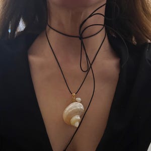 Puede incluir: Un collar de cordón negro largo con un colgante de concha marina espiral natural y un pequeño acento de perla blanca. El cordón está atado en un nudo suelto alrededor del cuello, con la concha y la perla suspendidas de una anilla dorada.