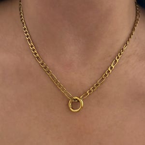 May include: A gold-toned figaro chain necklace featuring a series of flattened oval links interspersed with smaller circular links. The chain is secured at the center with a prominent, smooth, circular ring pendant that connects both ends of the chain.