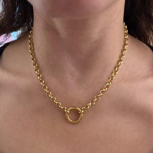 May include: A gold-colored chain necklace with a large circular pendant. The necklace features a chunky chain design with a polished finish. The pendant is a simple, textured circle. The necklace is worn around the neck.