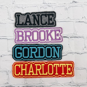 May include: Four embroidered name patches: "LANCE" in gray, "BROOKE" in purple, "GORDON" in teal, and "CHARLOTTE" in red. Each patch has a contrasting border and is displayed against a white brick wall.