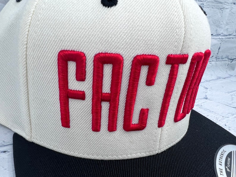K&ouml;nnte beinhalten: Wei&szlig;e und schwarze Baseballkappe mit rotem Sticktext "FACTOR".