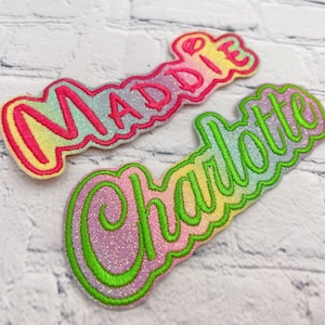 May include: Two embroidered name patches, "Maddie" and "Charlotte", in a playful, cursive font. "Maddie" features pink and yellow gradient lettering with a pink outline. "Charlotte" has green lettering with a green outline, set against a glittery background.
