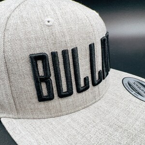K&ouml;nnte beinhalten: Eine graue und schwarze Baseballkappe mit dem Wort "BULLA" in schwarzer Stickerei auf der Vorderseite. Die Kappe hat einen gebogenen Schirm und einen Druckknopfverschluss.
