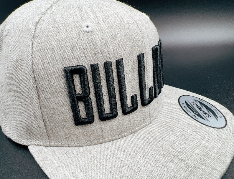 K&ouml;nnte beinhalten: Eine graue und schwarze Baseballkappe mit dem Wort "BULLA" in schwarzer Stickerei auf der Vorderseite. Die Kappe hat einen gebogenen Schirm und einen Druckknopfverschluss.