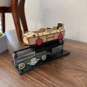 May include: A brown and tan model of a mine train ride with red wheels. The model is on a black base with a silver track and a black and green sign that reads "The Black  Mine Cars".
