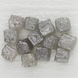 Puede incluir: Un grupo de pequeños diamantes grises de forma cuadrada. Los diamantes son en bruto y tienen un aspecto natural sin pulir.