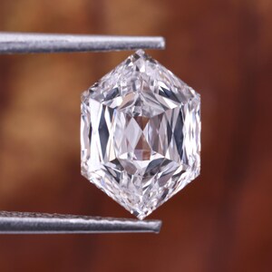 May include: A clear, hexagonal-cut diamond held by tweezers. The gemstone's facets reflect light, highlighting its intricate design. The background is a blurred, warm brown colour.