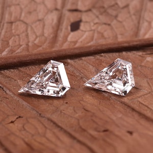 May include: Two clear, triangular-cut gemstones with multiple facets, reflecting light. The gems are set against a textured, brown background, possibly a leaf. The stones are likely diamonds or similar precious stones.