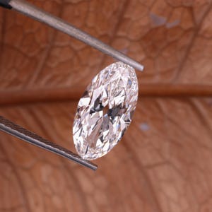 May include: An oval-cut diamond, held by tweezers, with multiple facets reflecting light. The diamond appears clear and bright. The background is a blurred, warm brown colour.
