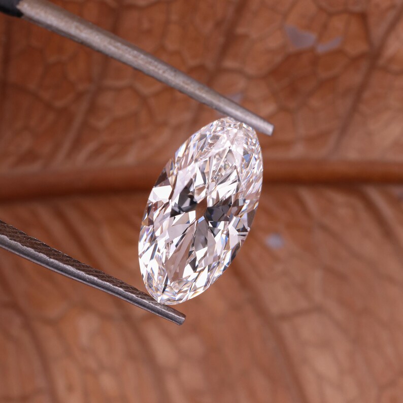 May include: An oval-cut diamond, held by tweezers, with multiple facets reflecting light. The diamond appears clear and bright. The background is a blurred, warm brown colour.
