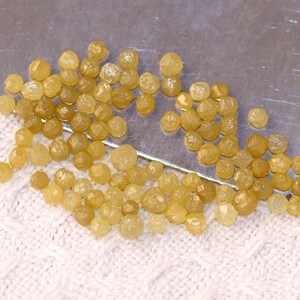 May include: A collection of small, yellow, rough diamonds, also known as uncut diamonds, scattered on a white surface. The diamonds are various sizes and shapes.