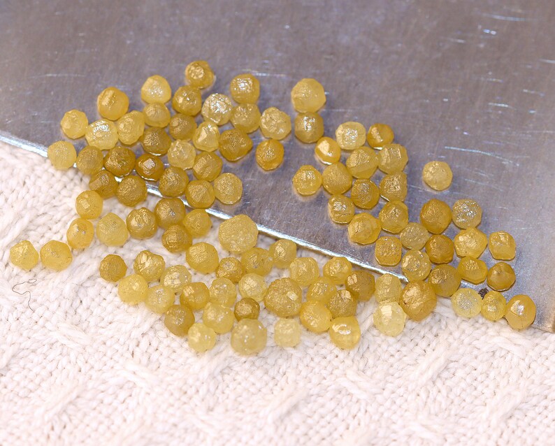 May include: A collection of small, yellow, rough diamonds, also known as uncut diamonds, scattered on a white surface. The diamonds are various sizes and shapes.