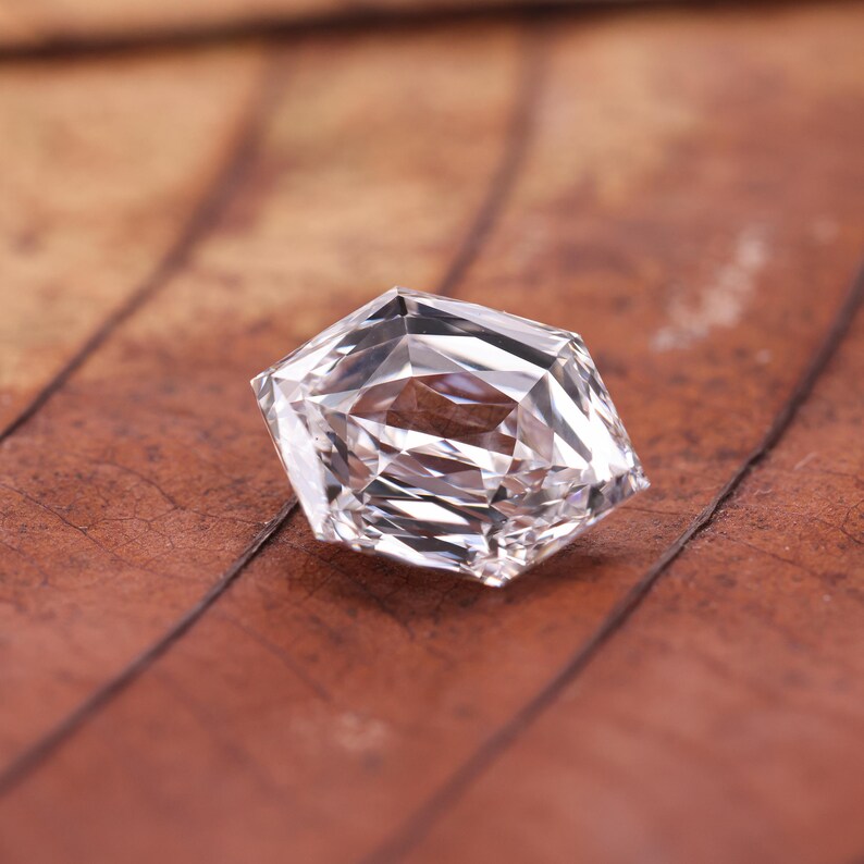 May include: A faceted, clear gemstone with a unique geometric shape. The gem is set against a brown, textured background, highlighting its brilliance and clarity. The cut of the stone creates multiple reflective surfaces.