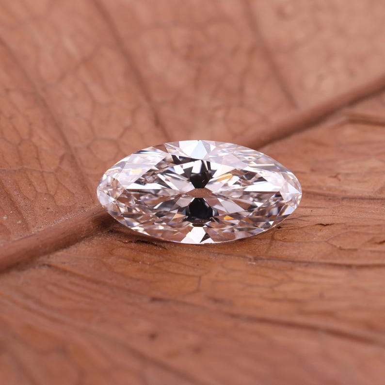 May include: A clear marquise-cut diamond, displaying multiple facets that catch the light. The gemstone sits on a brown, textured surface. The elongated shape of the diamond is a key feature, highlighting its sparkle. The image showcases a piece of jewelry, a gemstone.