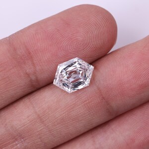 May include: A clear, hexagonal-cut diamond with multiple facets, reflecting light. The gemstone is held in a person's hand, showcasing its brilliance and clarity. The diamond's shape is a unique, modern design, perfect for jewelry.