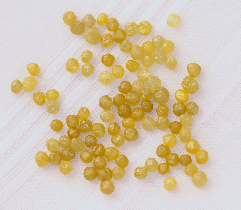 May include: A collection of small, yellow, faceted gemstones, likely diamonds, scattered on a white fabric surface.