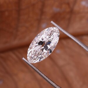 May include: A marquise-cut diamond held by tweezers. The clear, faceted diamond reflects light. The background is a blurred brown.
