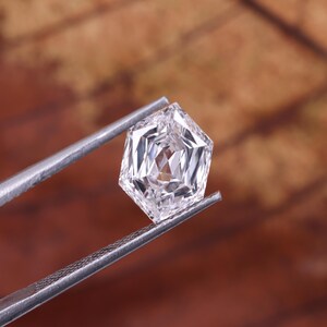 May include: A faceted, clear gemstone held by silver tweezers. The gemstone has a unique hexagonal shape with multiple facets, reflecting light. The background is a blurred, warm brown.