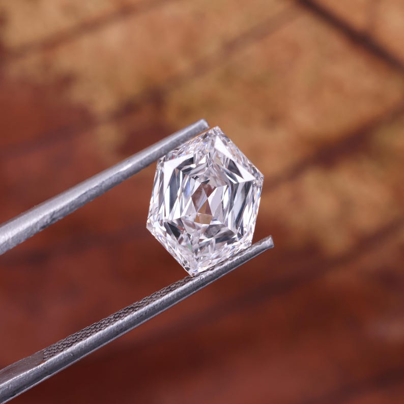 May include: A faceted, clear gemstone held by silver tweezers. The gemstone has a unique hexagonal shape with multiple facets, reflecting light. The background is a blurred, warm brown.