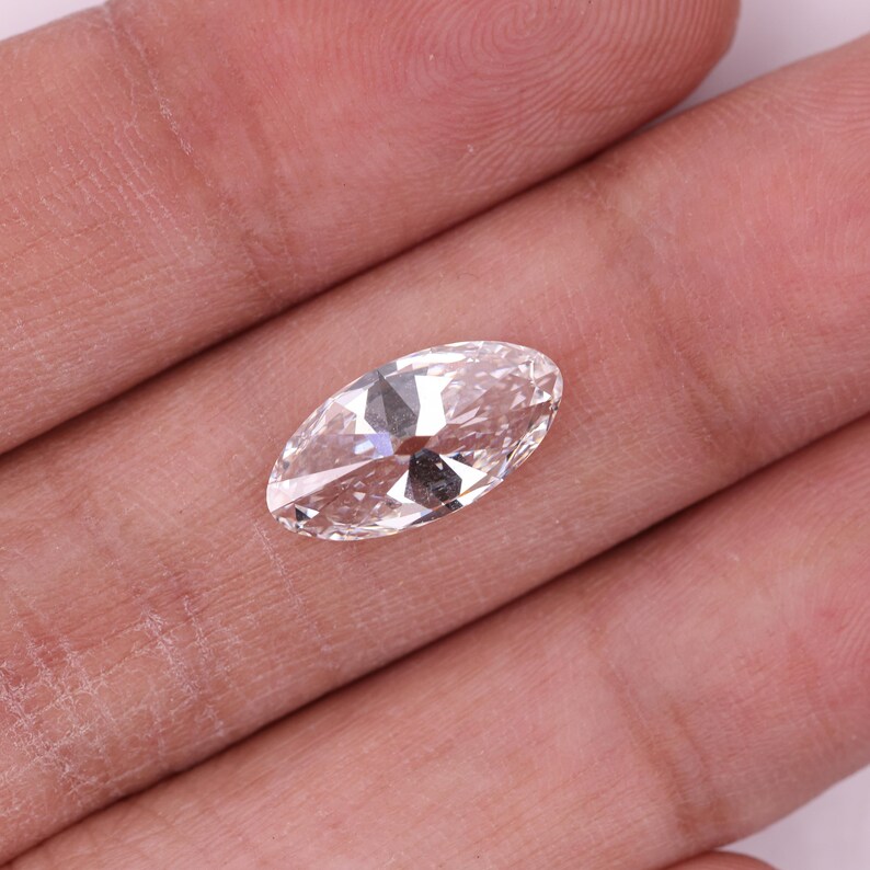 May include: A clear, oval gemstone with multiple facets, held in a hand. The gem reflects light, showcasing its cut and clarity. The stone appears to be colourless, with a brilliant sparkle.