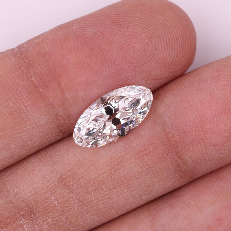 May include: A clear, oval-cut diamond with multiple facets, held in a hand. The diamond reflects light, showcasing its brilliance. The gem is set with small metal prongs. The background is a light pink.