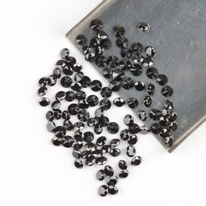 May include: A large quantity of black round gemstones, likely diamonds, are scattered on a white surface. Some of the gemstones are spilling from a metal tray.