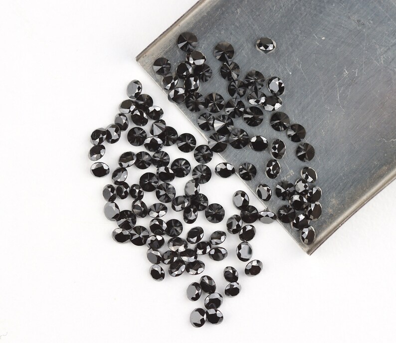 May include: A large quantity of black round gemstones, likely diamonds, are scattered on a white surface. Some of the gemstones are spilling from a metal tray.