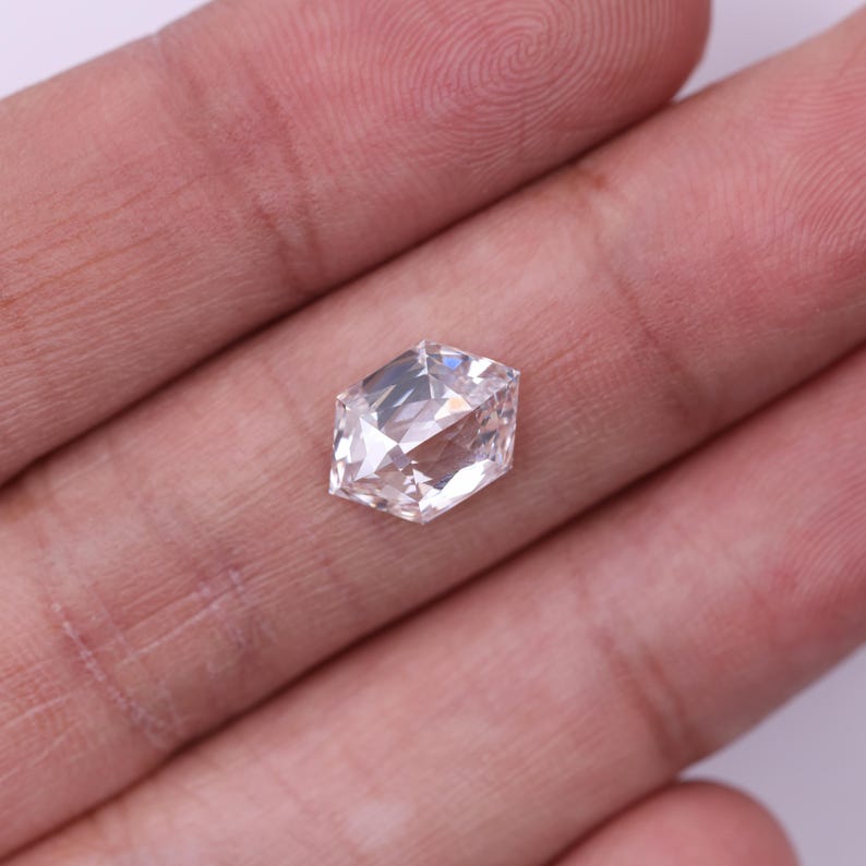 May include: A faceted, colorless gemstone with a hexagonal shape. The gem is held in a hand, showcasing its multiple reflective surfaces and brilliant sparkle. The stone appears to be a diamond, cut to maximize light reflection.