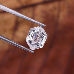 May include: A faceted, clear gemstone held by silver tweezers. The gem has a unique, geometric cut with multiple facets, reflecting light. The background is a blurred, warm brown.