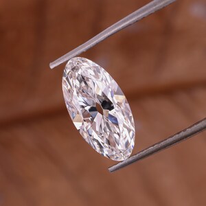 May include: A marquise-cut diamond held by tweezers. The diamond is clear and faceted, reflecting light. The shape is elongated with pointed ends. The background is a warm brown.
