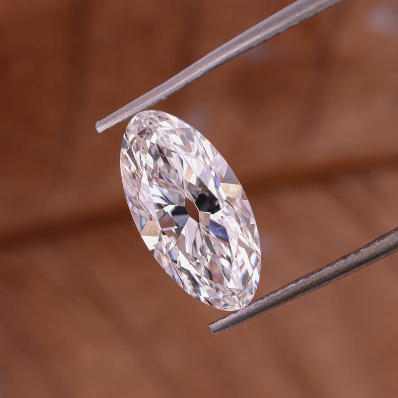 May include: A marquise-cut diamond held by tweezers. The diamond is clear and faceted, reflecting light. The shape is elongated with pointed ends. The background is a warm brown.
