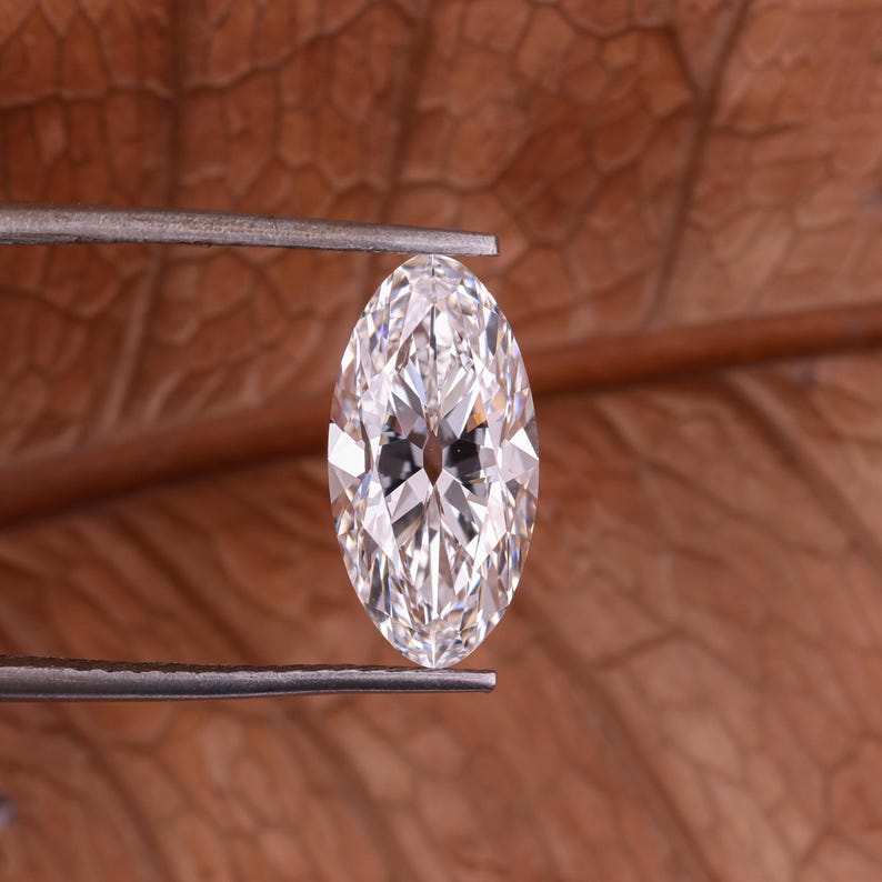 May include: A marquise-cut diamond held by tweezers. The diamond is clear and faceted, reflecting light. The background is a blurred, brown, textured surface.
