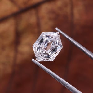 May include: A faceted, hexagonal-cut diamond held by silver tweezers. The gemstone is clear and reflects light, showcasing its internal facets. The background is a blurred brown.