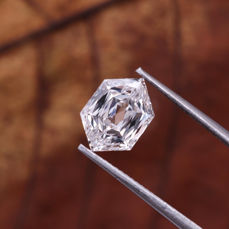 May include: A faceted, hexagonal-cut diamond held by silver tweezers. The gemstone is clear and reflects light, showcasing its internal facets. The background is a blurred brown.