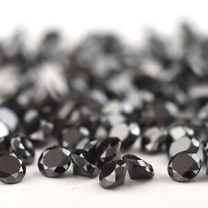May include: A group of black round gemstones, likely diamonds, scattered on a white surface. The gemstones are faceted and reflect light.