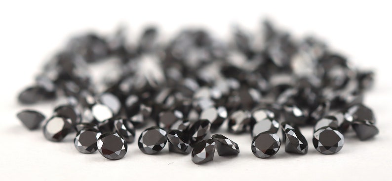 May include: A group of black round gemstones, likely diamonds, scattered on a white surface. The gemstones are faceted and reflect light.