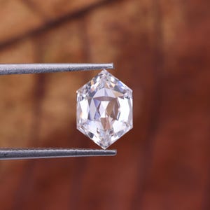 May include: A clear, hexagonal-cut gemstone held by tweezers. The gem has multiple facets that reflect light, creating a brilliant sparkle. The background is a blurred, warm brown.