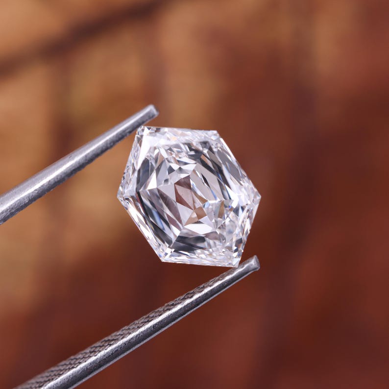 May include: A faceted, clear diamond held by silver tweezers. The diamond has a unique hexagonal shape with multiple facets, reflecting light. The background is a blurred brown.