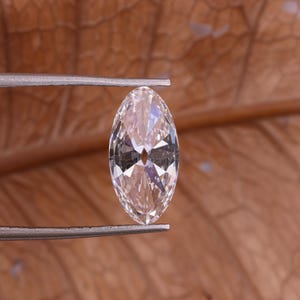 May include: A marquise-cut gemstone, held by tweezers, shows a clear, faceted surface. The gem has a light, warm hue and is set against a blurred, brown background. The elongated shape and multiple facets create a sparkling effect.