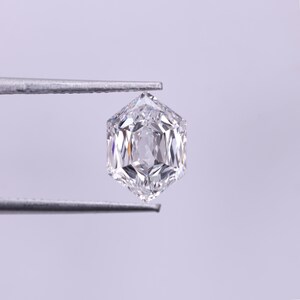 May include: A clear, hexagonal-cut diamond held by silver tweezers. The diamond has multiple facets that reflect light, creating a brilliant sparkle. The gemstone is set against a soft, light purple background.