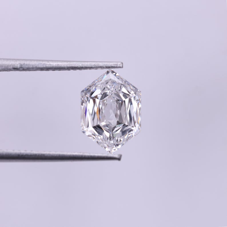 May include: A clear, hexagonal-cut diamond held by silver tweezers. The diamond has multiple facets that reflect light, creating a brilliant sparkle. The gemstone is set against a soft, light purple background.