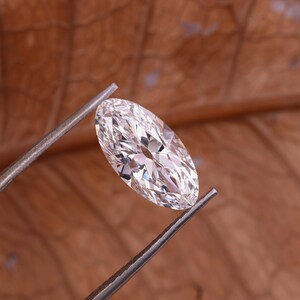 May include: A clear, oval-cut diamond held by metal tweezers. The gem is highly faceted, reflecting light. The background is a warm, brown tone, providing contrast to the brilliant stone. The diamond is a classic shape, often used in engagement rings.