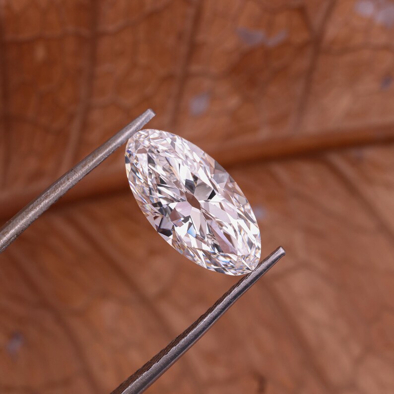 May include: A clear, oval-cut diamond held by metal tweezers. The gem is highly faceted, reflecting light. The background is a warm, brown tone, providing contrast to the brilliant stone. The diamond is a classic shape, often used in engagement rings.
