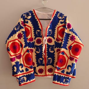 Puede incluir: Una chaqueta de color crema con cuello en V y mangas largas. La chaqueta está bordada con un patrón geométrico en tonos de azul, rojo y rosa.